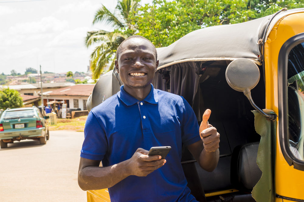 cheerful african man standing next to his tuk tuk keke taxi smiling and using his smart phone giving a thumbs up